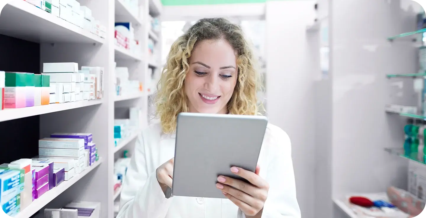 Smiling pharmacist reading a tablet between pharmacy shelves